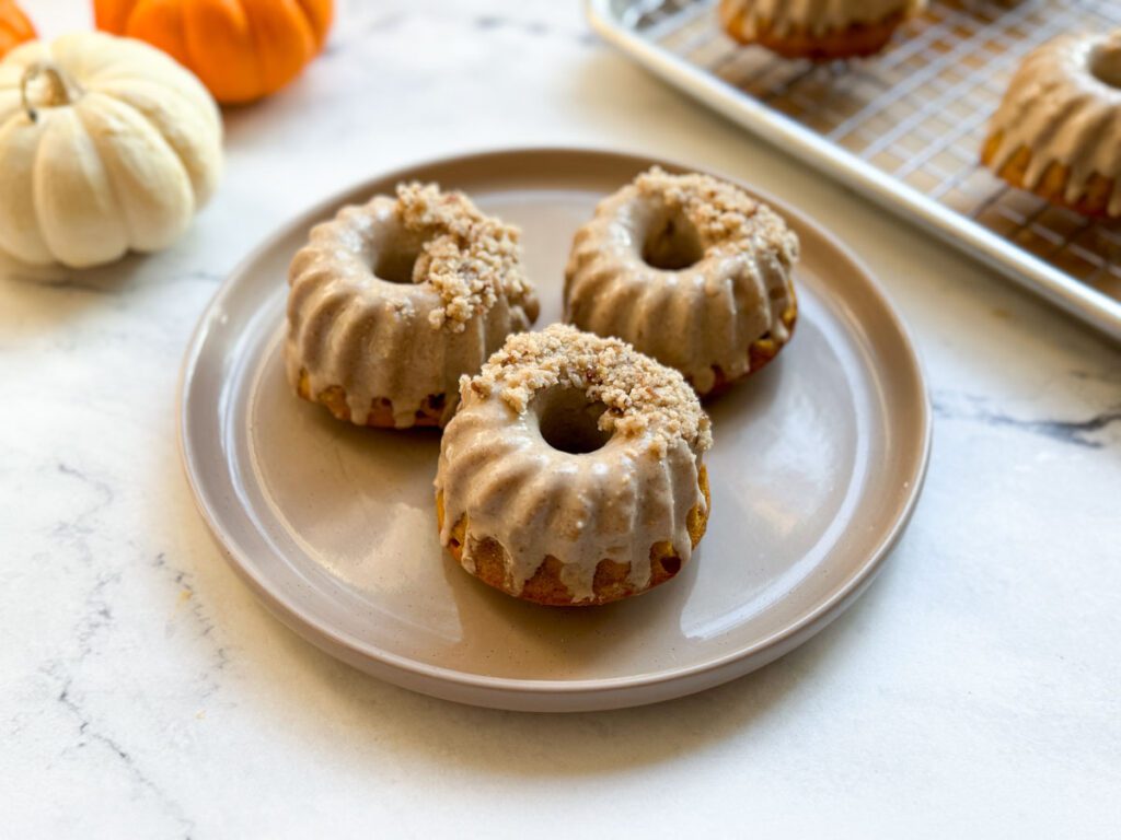 gluten-free pumpkin donuts on plate with mini pumpkins and additional donuts on baking tray in background
