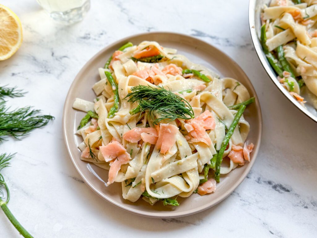plated smoked salmon and asparagus pasta with fresh dill, lemon, and pan of pasta visible in the background